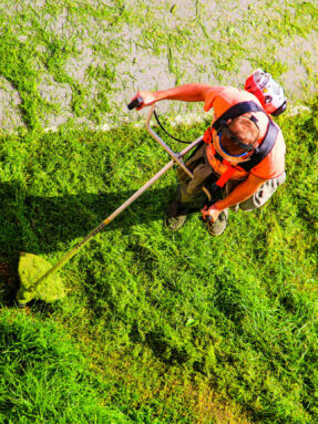 Young worker mowing lawn with grass trimmer outdoors on sunny day. Man mowing the grass, the mower top view. Professional roadside mowing. Photo of young man mowing the grass during beautiful evening